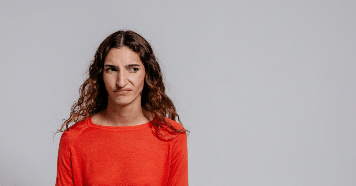 June Blog Images 12 - San Jose Mental Health Woman with curly brown hair in an orange sweater looks to the left with a puzzled expression against a gray backdrop.