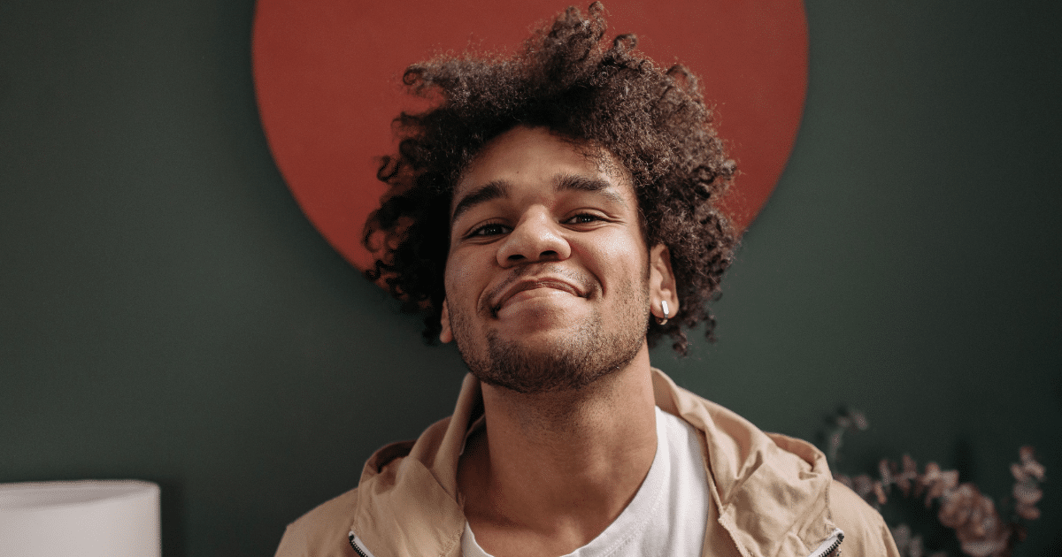June Blog Images 13 - San Jose Mental Health Young man with curly hair and an earring, wearing a beige jacket over a white shirt, smiles at the camera against a dark green wall with a red circle behind him.