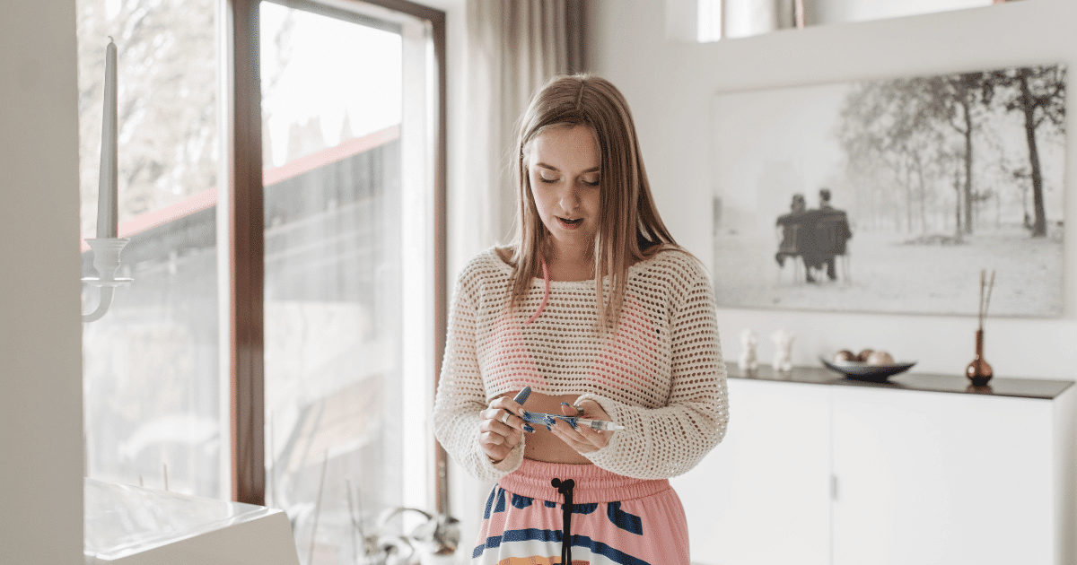 June Blog Images 18 - San Jose Mental Health Young woman in a white knit sweater and pink striped shorts stands in a bright living room, looking at her smartphone.