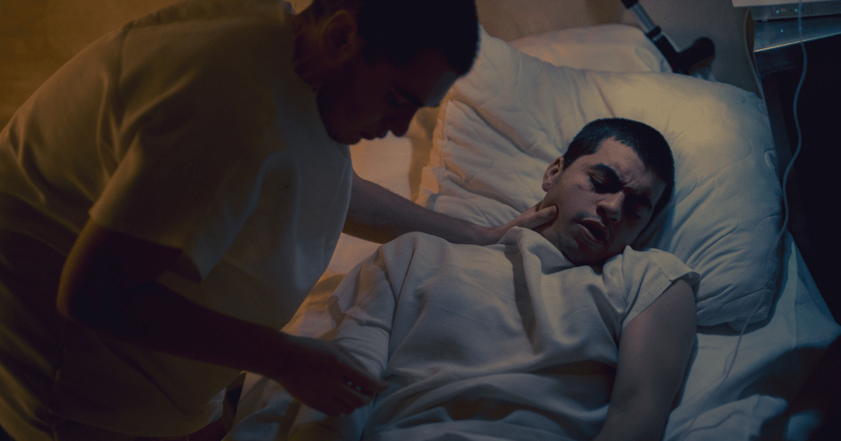 JULY-BLOG IMAGES - San Jose Mental Health Caregiver tending to a patient lying in a hospital bed, supporting his neck in a dimly lit room with monitoring equipment nearby