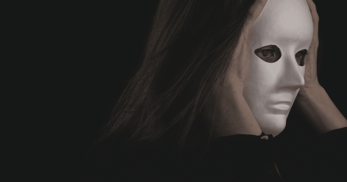 JULY-BLOG IMAGES 69 - San Jose Mental Health Person with long hair holds a white blank mask up to their face, hiding their identity against a dark background.