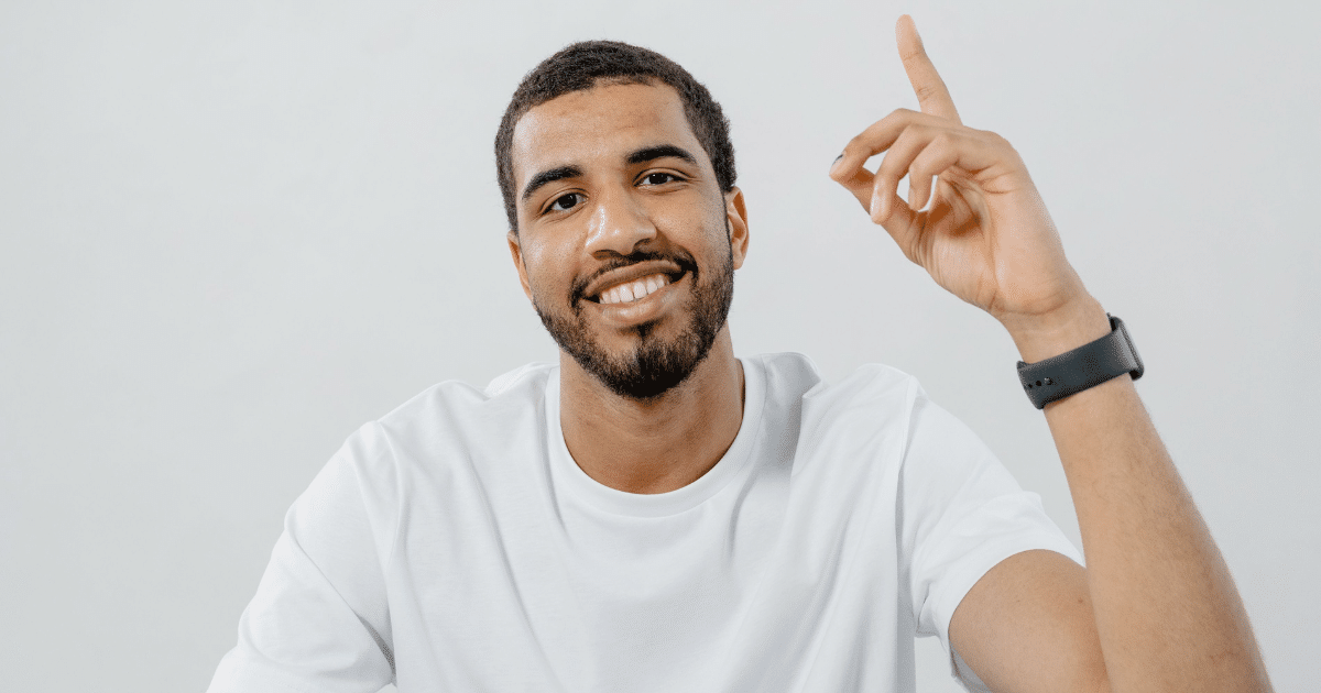 August-Blog Images - San Jose Mental Health Smiling man in a white T-shirt raises his index finger as if making a point, wearing a black smartwatch.